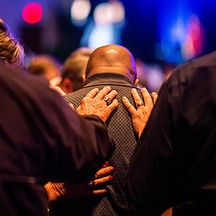Close-up of hands on shoulders in prayer with stage lights in the background