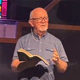 Dr. Marrow holds an open Bible while preaching, with stained glass in the background